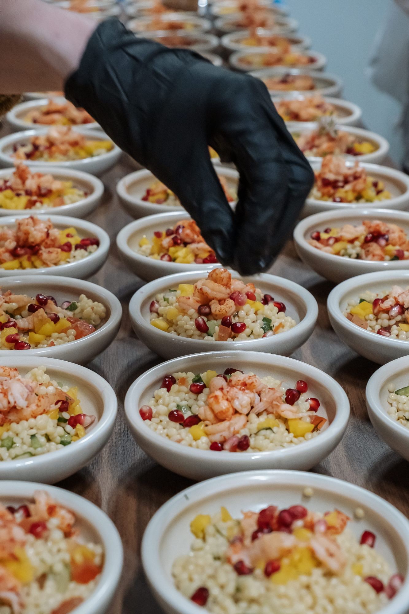 Een lange tafel vol kommetjes parelcouscous met scampi en granaatappel, een hand in een zwarte handschoen werkt ze af